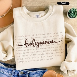 May include: Sand-colored sweatshirt with the word "holyween" in a cursive font. The sweatshirt has a crew neck and long sleeves. Text on the shirt describes a Christian take on Halloween. A brown hat, denim shorts, and a small succulent are also in the image.