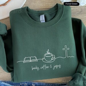 May include: Forest green sweatshirt with a white line drawing of an open book, coffee cup, and cross. The text "books, coffee & jesus" is below the drawing. The sweatshirt is folded and has a crew neck.