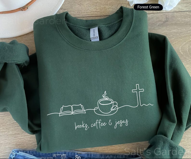 May include: Forest green sweatshirt with a white line drawing of an open book, coffee cup, and cross. The text "books, coffee & jesus" is below the drawing. The sweatshirt is folded and has a crew neck.