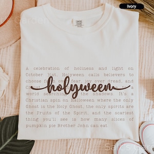 May include: Ivory-colored t-shirt with the word "holyween" in a cursive font. The shirt features text describing a Christian take on Halloween. The shirt is part of the Comfort Colors collection.