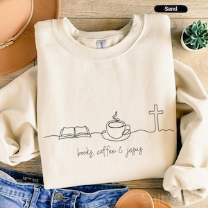May include: Sand-colored sweatshirt with a minimalist black line drawing of an open book, coffee cup, and cross. The words "books, coffee & jesus" are written in a handwritten font. The sweatshirt is folded on a wooden surface.
