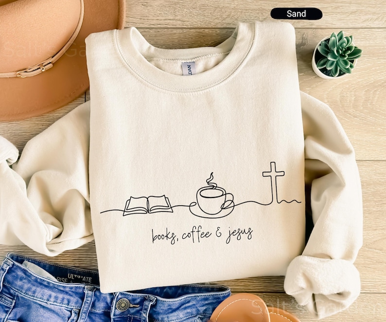 May include: Sand-colored sweatshirt with a minimalist black line drawing of an open book, coffee cup, and cross. The words "books, coffee & jesus" are written in a handwritten font. The sweatshirt is folded on a wooden surface.