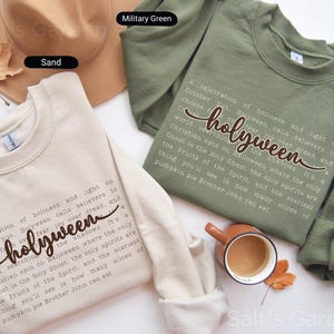 K&ouml;nnte beinhalten: Zwei Sweatshirts mit Rundhalsausschnitt, eines in Sand und eines in Milit&auml;rgr&uuml;n, werden gezeigt. Das sandfarbene Sweatshirt hat den Text "holyween" in einer Schreibschrift, darunter ein Gedicht. Das gr&uuml;ne Sweatshirt hat das gleiche Design. Ein brauner Hut und eine Tasse Kaffee sind ebenfalls im Bild.