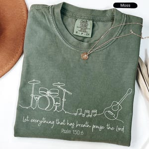 May include: Moss green t-shirt with a white line drawing of a drum set, guitar, and musical notes. The text "Let everything that has breath praise the Lord Psalm 150:6" is printed below the drawing. A gold necklace is draped on the shirt.