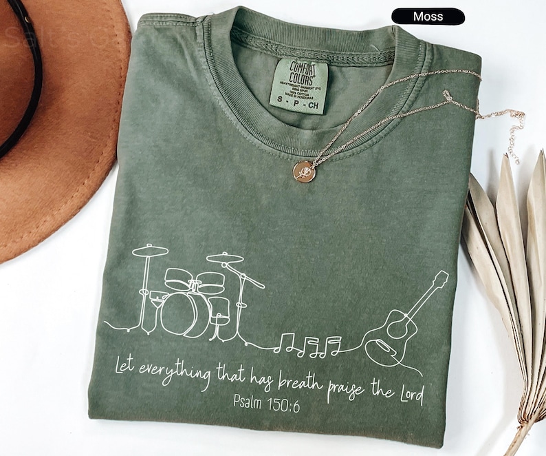 May include: Moss green t-shirt with a white line drawing of a drum set, guitar, and musical notes. The text "Let everything that has breath praise the Lord Psalm 150:6" is printed below the drawing. A gold necklace is draped on the shirt.