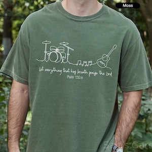 May include: Moss green t-shirt with a white line drawing of a drum set, musical notes, and a guitar. The text "Let everything that has breath praise the Lord Psalm 150:6" is printed below the drawing. The shirt is a comfortable fit.