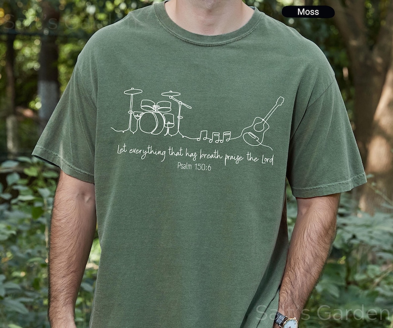 May include: Moss green t-shirt with a white line drawing of a drum set, musical notes, and a guitar. The text "Let everything that has breath praise the Lord Psalm 150:6" is printed below the drawing. The shirt is a comfortable fit.