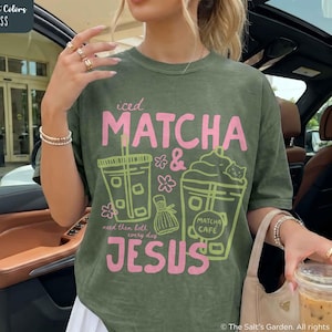 May include: A moss green t-shirt featuring a graphic print with the text "iced MATCHA & JESUS" in pink and "need them both every day" in smaller pink font. The design includes line art of two drinks, a small whisk, and pink flowers.