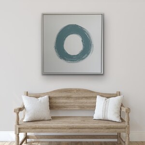 May include: A framed abstract painting with a teal blue circle on a white background hangs above a wooden bench. The bench has two white cushions with striped patterns. The room has light wood flooring and a white rug.