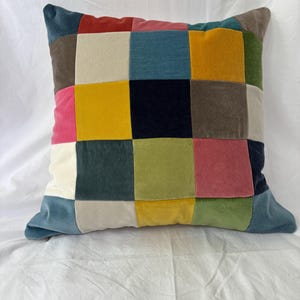 Peut inclure: Coussin carré décoratif avec un motif patchwork. Le coussin présente une grille de carrés de différentes couleurs, dont le rouge, le rose, le jaune, le bleu, le vert et le marron. Le coussin est en matière douce, semblable au velours.