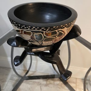 May include: A black wooden bowl with a wide rim and a decorative exterior featuring a mosaic-like pattern of dark, earthy-toned shapes. The bowl is supported by a black wooden stand with three distinct, angled legs that provide a stable base.