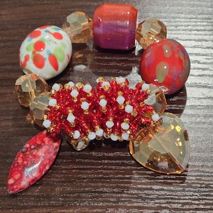 Puede incluir: Una pulsera de cuentas con una variedad de cuentas coloridas. La pulsera incluye cuentas rojas, blancas y doradas, junto con cuentas más grandes en tonos de rojo, naranja y transparente. La pulsera se muestra sobre una superficie de madera oscura.