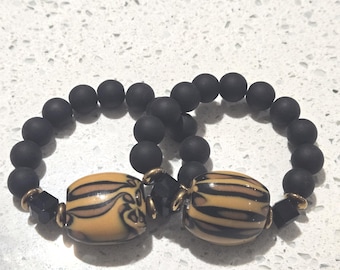 May include: Two beaded bracelets. Each bracelet features matte black round beads and a central oval bead with a tan and black striped design. The bracelets are connected with gold-toned metal and black geometric beads.