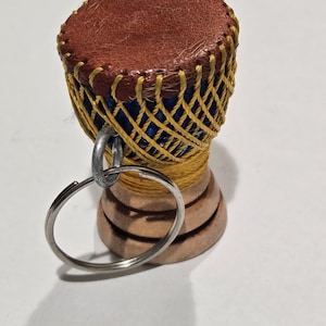May include: Miniature drum keychain with a brown leather top and a woven yellow and blue body. The base is made of stacked wooden rings. A silver key ring is attached.