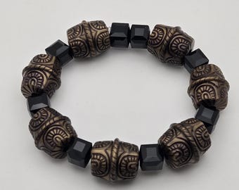 May include: A beaded bracelet featuring alternating dark bronze-colored, intricately patterned beads and black, square-cut beads. The bracelet is set against a plain white background, highlighting the contrast of colors and textures.