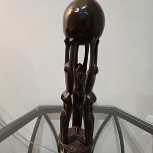 May include: A dark brown wooden sculpture featuring a large sphere held aloft by three stylized figures. The figures are carved with simple, elegant lines, and the base has a textured pattern. The sculpture is displayed on a glass table.