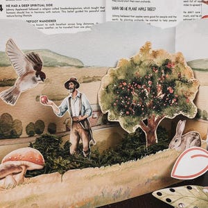 May include: A three-dimensional paper diorama featuring a bearded figure in a hat and suspenders holding an apple, a large apple tree, a flying bird, a rabbit, and a mushroom. The scene is set against a printed background with text.