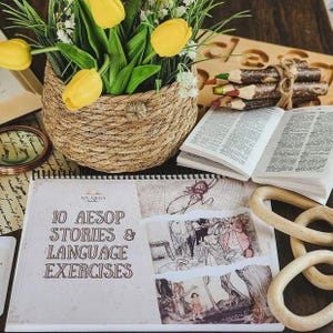 Op de afbeelding: Een spiraalgebonden boek getiteld 10 Aesop Stories & Language Exercises met vintage illustraties, een gevlochten mand met gele tulpen, een bundel rustieke kleurpotloden in takvorm, een open boek en drie houten ringen.