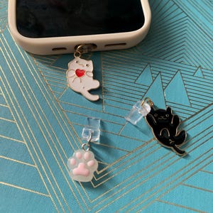 Puede incluir: Tres charms para teléfono con un gato blanco sosteniendo un corazón rojo, un gato negro y una huella de pata rosa. Los charms están unidos a soportes de plástico transparente.