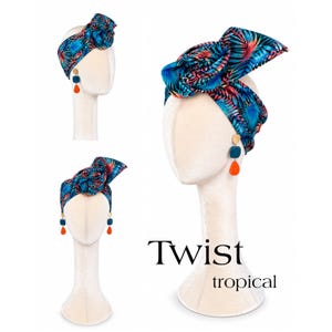 Può includere: Una fascia per capelli con motivo a foglie tropicali nei toni del blu, nero e rosa, annodata con un nodo superiore prominente. La fascia è esposta su una testa di manichino insieme a orecchini pendenti a forma di goccia color ottanio e arancione. Il testo "Twist tropical" è visibile.