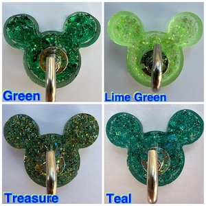 May include: Four resin wall hooks shaped like mouse heads, each featuring a metal hook and glittery finish. The colors are labeled as Green, Lime Green, Treasure, and Teal, with each hook displaying distinct shades of green and gold glitter.