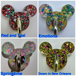 May include: Four resin badge reels shaped like mouse heads, each featuring a metal clip and colorful glitter. The designs are labeled Red and Teal, Emotions, Springtime, and Down in New Orleans, showcasing various glitter color combinations.