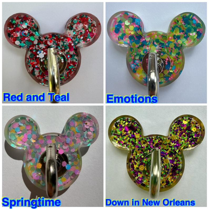 May include: Four resin badge reels shaped like mouse heads, each featuring a metal clip and colorful glitter. The designs are labeled Red and Teal, Emotions, Springtime, and Down in New Orleans, showcasing various glitter color combinations.