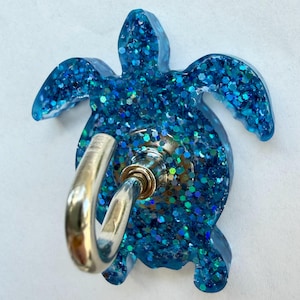 Könnte beinhalten: Ein blau glitzernder, schildkrötenförmiger Telefonhalter mit einem silbernen Metallring. Die Schildkröte hat eine glänzende, strukturierte Oberfläche mit eingebetteten Glitzerpartikeln. Der Ring ist mit einem Drehmechanismus an der Rückseite befestigt.