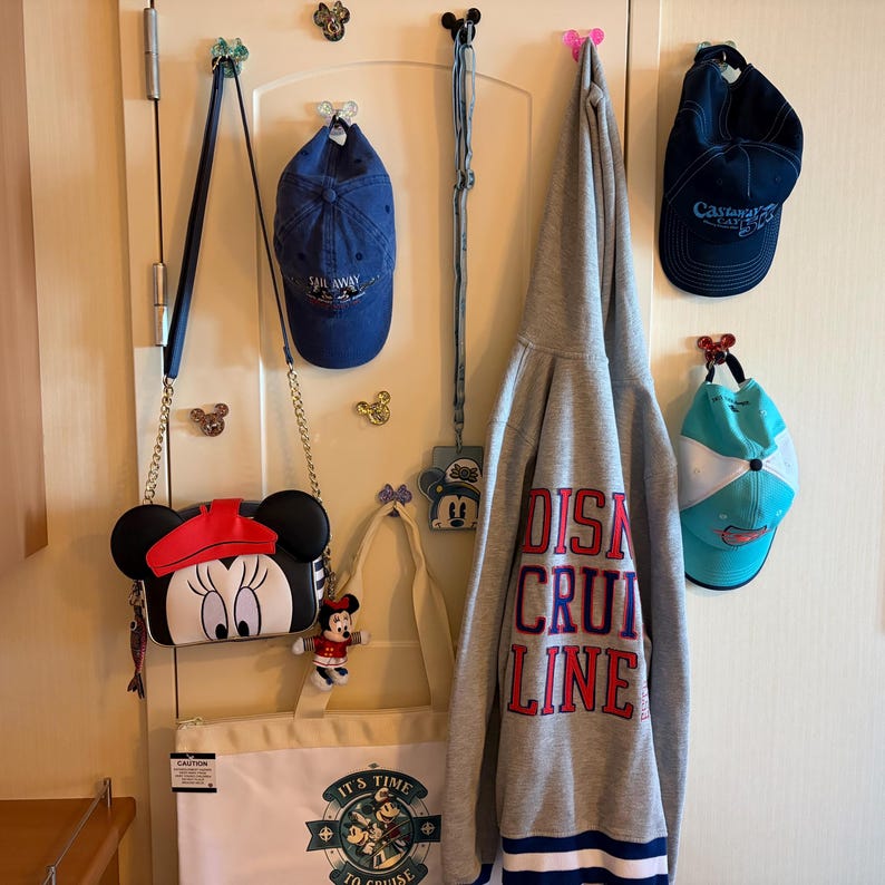 May include: A collection of items hanging on a door including a blue baseball cap, a grey hoodie with Disney Cruise Line text, a white tote bag with a cruise graphic, a Minnie Mouse crossbody bag, a lanyard, and a teal and white cap.