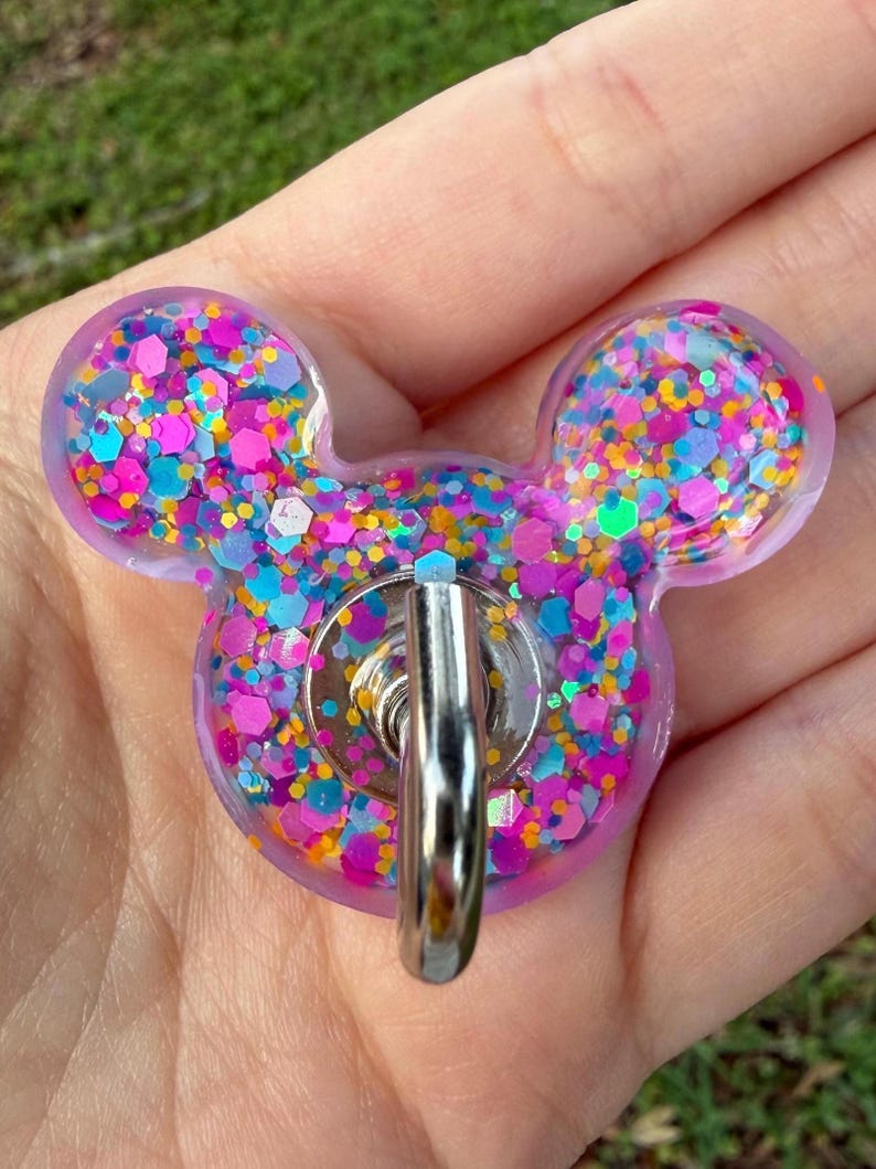 May include: A mouse-shaped resin badge reel featuring a translucent purple base filled with a dense mix of multicolored hexagonal and circular glitter in shades of pink, blue, orange, and silver, with a central silver metal belt clip.