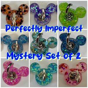 Puede incluir: Un conjunto de soportes para teléfono con forma de Mickey Mouse en varios colores: naranja, morado, verde, azul y rosa. Cada soporte está lleno de purpurina y tiene un anillo plateado. El texto "Perfectly Imperfect Mystery Set of 2" está presente.