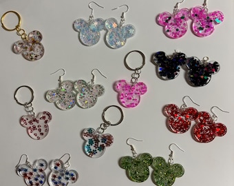 May include: A collection of resin accessories shaped like mouse heads, including several pairs of dangle earrings with silver-toned hooks and keychains with gold or silver-toned rings, featuring various glitter, floral, and star-shaped inserts.