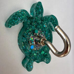 Op de afbeelding: Een wandhaak in de vorm van een zeeschildpad, gemaakt van doorschijnende blauwgroene hars met daarin verschillende maten groene, blauwe en iriserende glitters. Een zilverkleurige metalen haak is in het midden van het schild bevestigd.