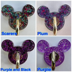 May include: Four resin badge reels shaped like mouse heads, each featuring a metal clip. The top left is labeled Scarers with multi-colored glitter, top right is Plum with purple and blue glitter, bottom left is Purple and Black, and bottom right is Purple.