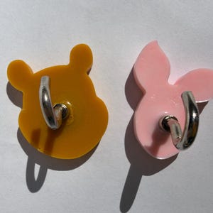May include: Two character-shaped phone grips. One is yellow and shaped like a bear's head, the other is pink and shaped like a pig's head. Both have silver metal finger rings attached to the back.