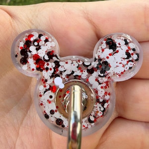 May include: A clear resin badge reel shaped like a mouse head, filled with a mix of white, black, and red circular glitter. A silver-toned metal belt clip is attached to the center of the resin piece, featuring a circular base and a hook.