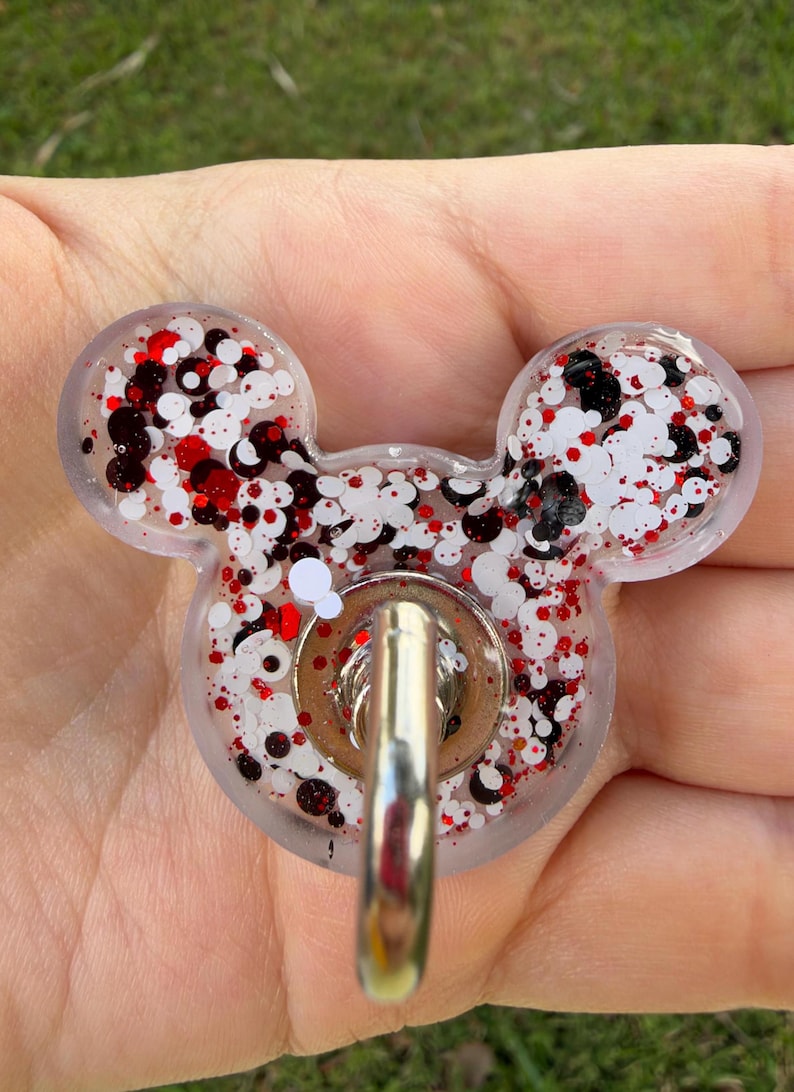 May include: A clear resin badge reel shaped like a mouse head, filled with a mix of white, black, and red circular glitter. A silver-toned metal belt clip is attached to the center of the resin piece, featuring a circular base and a hook.
