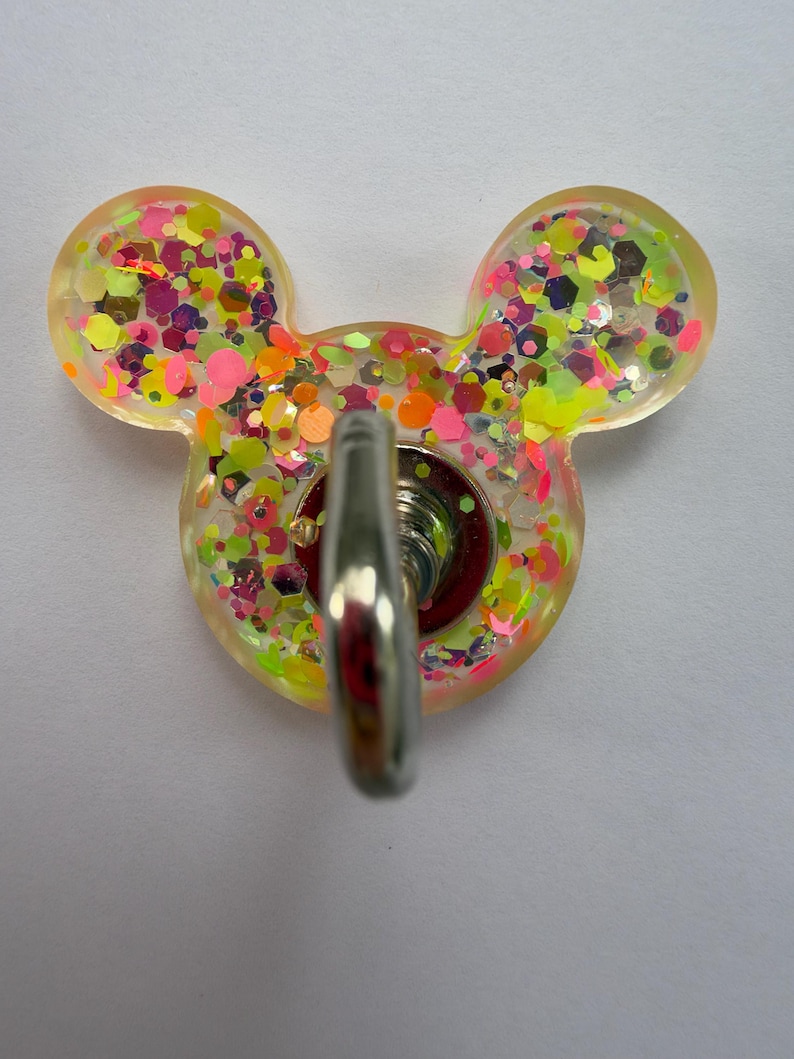 May include: A mouse-shaped wall hook made of translucent resin filled with multicolored neon glitter in shades of pink, yellow, and orange. The hook features a central silver-toned metal attachment point protruding from the center of the resin base.