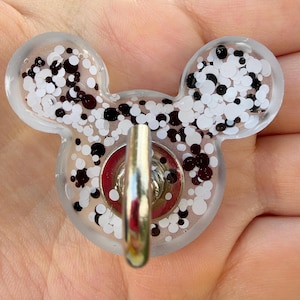 May include: A clear resin badge reel shaped like a mouse head, filled with a mix of white and black circular glitter pieces. A silver-toned metal hook is attached to the center of the resin piece, featuring a circular red base.