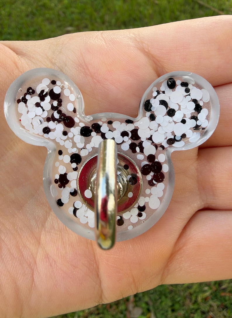 May include: A clear resin badge reel shaped like a mouse head, filled with a mix of white and black circular glitter pieces. A silver-toned metal hook is attached to the center of the resin piece, featuring a circular red base.