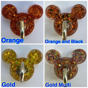 May include: Four resin wall hooks shaped like mouse heads, each featuring a metal hook. The designs include orange glitter, orange and black glitter, gold glitter, and a multi-colored glitter with thin metallic strips. Each hook is labeled.