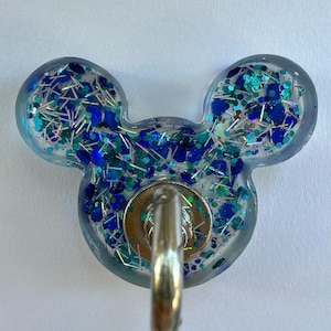 May include: A mouse-ear shaped resin badge reel featuring a clear base filled with blue, teal, and silver glitter in various shapes and sizes. A circular silver metal hardware component is attached to the center, featuring a retractable cord loop.