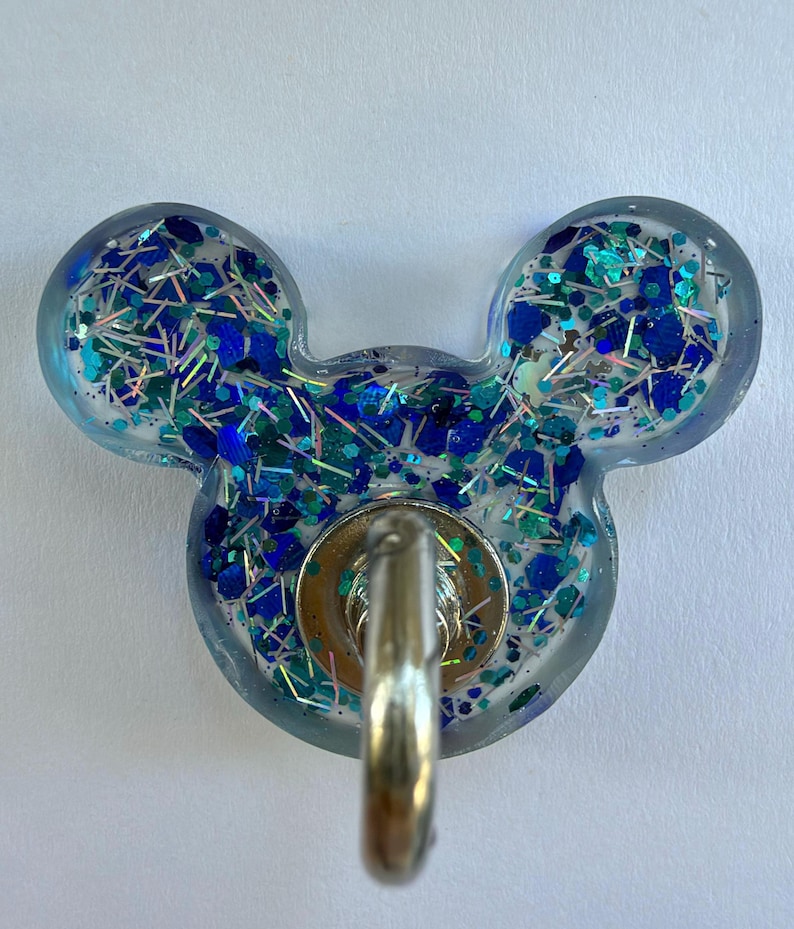 May include: A mouse-ear shaped resin badge reel featuring a clear base filled with blue, teal, and silver glitter in various shapes and sizes. A circular silver metal hardware component is attached to the center, featuring a retractable cord loop.