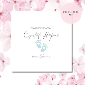 Puede incluir: Una tarjeta blanca con flores rosas alrededor de los bordes. La tarjeta dice "Diwrnod Mamau" y "Cyntaf Hapus" con una huella de pie azul con un corazón rosa en el centro. La tarjeta también dice "caru Oliver x".