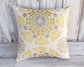 Puede incluir: Cojín cuadrado con fondo crema. Presenta un diseño floral bordado en amarillo y gris. El diseño es simétrico, con un motivo floral central y pequeños detalles florales en los bordes. El cojín se utiliza probablemente para la decoración del hogar, mide unos 40 cm x 40 cm.