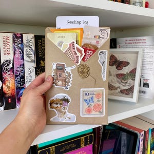 May include: A brown paper envelope containing a Reading Log card, decorated with various stickers including a classical bust, a stack of televisions, a postage stamp with flowers and a butterfly, and a gold wax seal on the front flap.