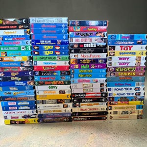 Puede incluir: Una gran colección de cintas VHS apiladas en tres columnas verticales. Los lomos muestran varios títulos de películas, incluidos Dead Man on Campus, Coyote Ugly, Caddyshack II, Bruce Almighty, Black Sheep, Black Knight y muchos otros en varios colores.