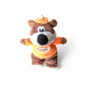 Pode incluir: Um urso de pelúcia marrom vestindo uma roupa laranja e marrom com o texto "A&W". O urso tem um focinho branco e um pequeno chapéu laranja. O bicho de pelúcia está em pé com os braços estendidos.