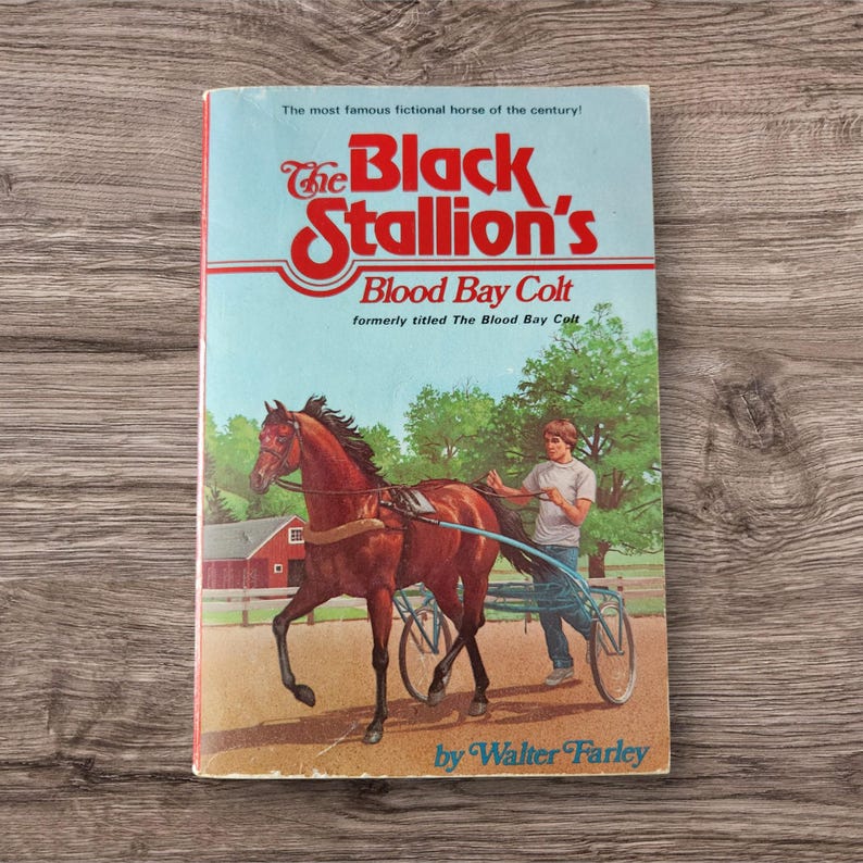 May include: A vintage paperback book titled "The Black Stallion's Blood Bay Colt" by Walter Farley. The cover features a brown horse pulling a blue cart, with a boy holding the reins. The background shows a red barn and green trees.