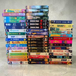 Könnte beinhalten: Eine große Sammlung von VHS-Kassetten, die in drei Spalten gestapelt sind. Die Buchrücken zeigen verschiedene Filmtitel wie The Lost World, Jurassic Park, Harry Potter, E.T., The Flintstones, Care Bears und andere mit bunten Grafikdesigns.